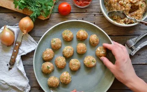 Polpette di pollo pronte per essere cucinate, disposte su un piatto grigio, con ingredienti freschi come cipolle e pomodoro sullo sfondo.