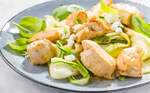 Succulento pollo e zucchine a spirale, guarnito con formaggio sbriciolato e basilico fresco, servito su un piatto blu.