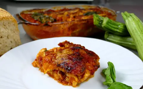 Una fetta di parmigiana di zucchine alla siciliana, dorata e succulenta, accanto a zucchine fresche e pane casereccio.