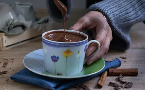 Una mano versa cioccolata fatta in casa in una tazza fiorata. Profumo di cannella e scaglie di cioccolato sul tavolo.