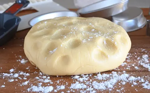 Impasto di pasta frolla con olio, pronto per essere lavorato. Si intravedono stampi per torte e utensili da cucina.