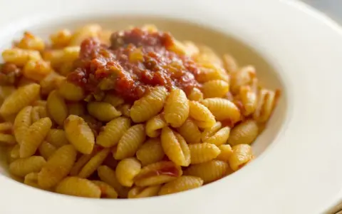 Malloreddus alla campidanese, pasta sarda con sugo di pomodoro e salsiccia, serviti in un piatto bianco.