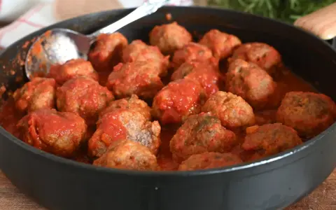 Un tegame pieno di polpette al sugo, pronte per essere gustate. Un cucchiaio d'argento è appoggiato sul bordo, invitando a servirle.
