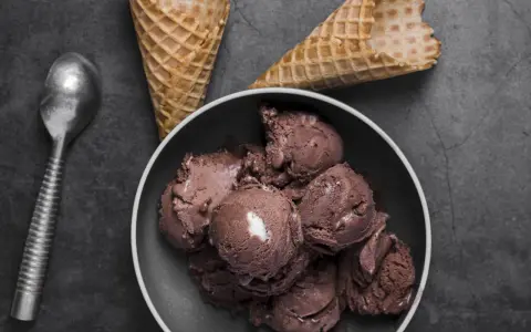 Coppa di **gelato al cioccolato** cremosa, con coni vuoti e un cucchiaio d'argento su sfondo scuro.