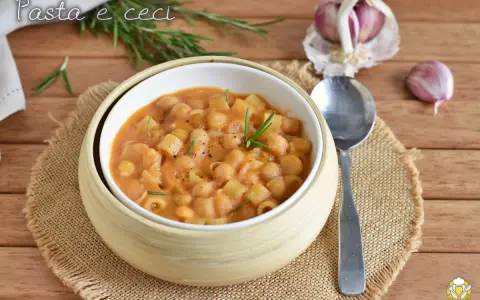 Un piatto fumante di pasta e ceci, guarnito con rosmarino fresco, su un tavolo di legno. Accanto, spicchi d'aglio e un cucchiaio.