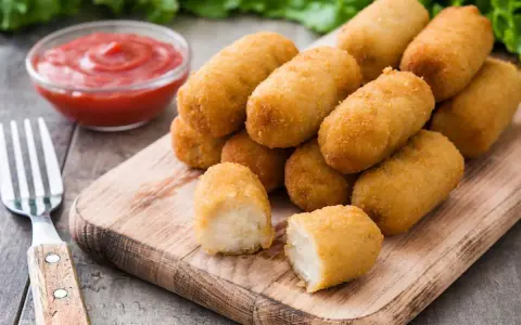 Crocchette di patate dorate e croccanti, servite su un tagliere di legno con ketchup e una forchetta.