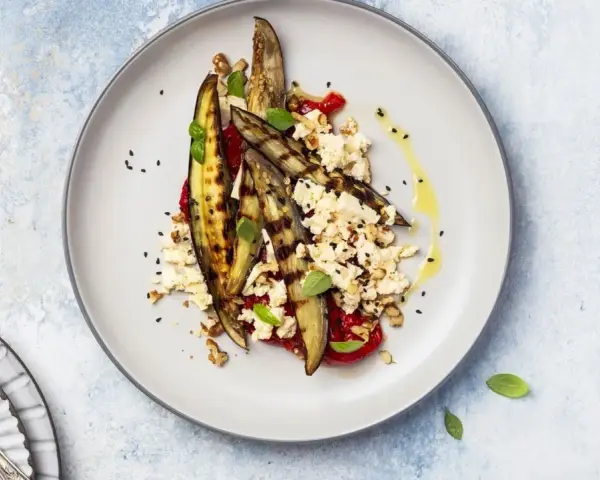 Deliziosa insalata di melanzane grigliate con feta sbriciolata, pomodoro, noci e semi di sesamo nero, condita con olio d'oliva.