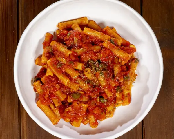 Un piatto di pasta alla pizzaiola, con rigatoni conditi da un sugo rosso ricco di pomodoro, capperi e origano, servito in una ciotola bianca su un tavolo di legno.