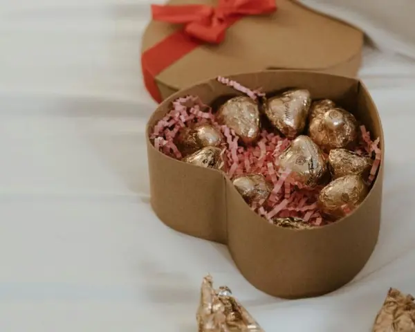 Scatola a forma di cuore con cioccolatini fatti in casa dorati su paglia rosa. Un'altra scatola a forma di cuore con fiocco rosso è sullo sfondo.