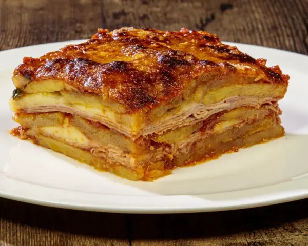 Una fetta di parmigiana di patate, stratificata con formaggio e salumi, servita su un piatto bianco.