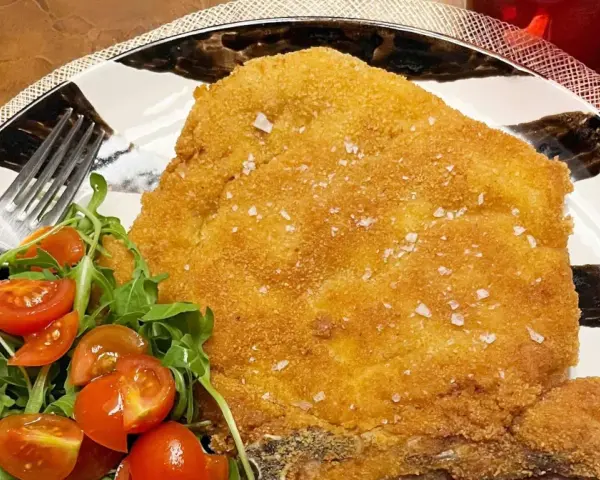 Una dorata cotoletta alla milanese, croccante e cosparsa di sale grosso, servita con un'insalata fresca di rucola e pomodorini.