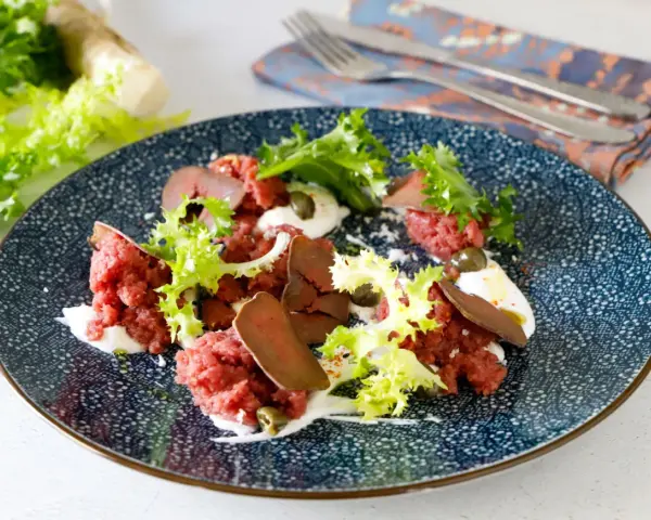 Deliziosa tartare di manzo con lattuga croccante, capperi e una crema bianca su un piatto blu decorato.