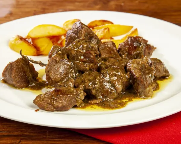 Un saporito abbacchio alla cacciatora, tenero e succulento, servito con patate arrosto su un piatto bianco.