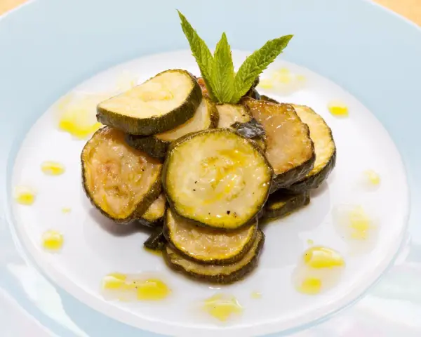 Fette di zucchine alla scapece, condite con olio e guarnite con menta fresca, su un piatto bianco.