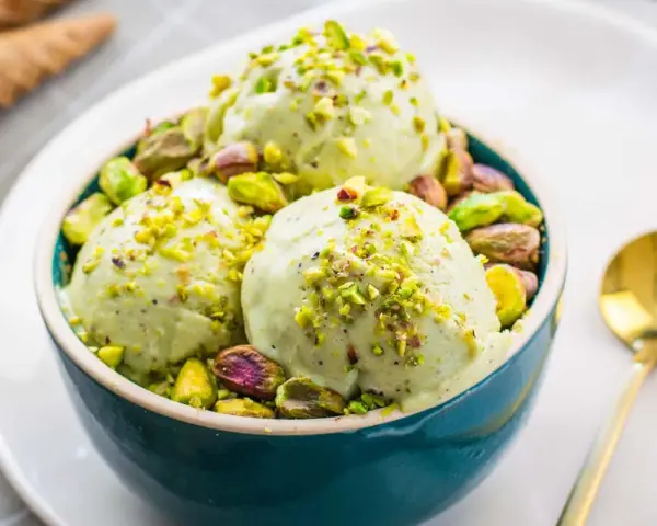 Tre palline di cremoso gelato al pistacchio, guarnite con granella e pistacchi interi, in una ciotola blu.