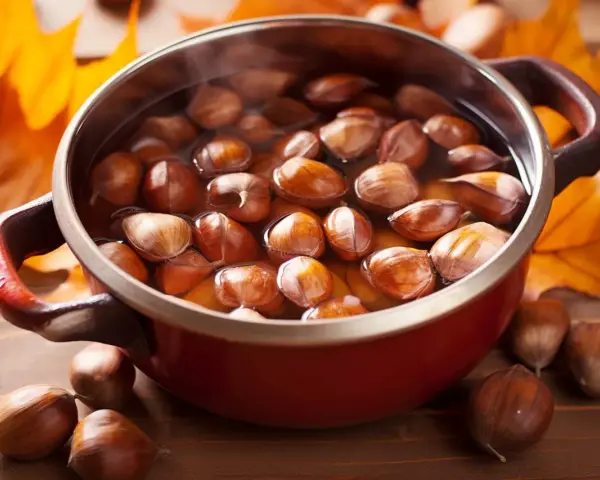 Castagne bollite con buccia in una pentola rossa, circondate da foglie autunnali.