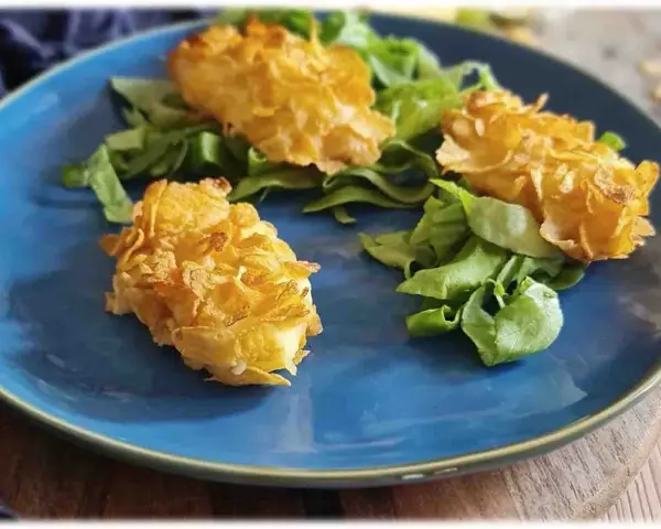 Tre croccanti polpette di patate impanate con fiocchi di mais, servite su un letto di insalata fresca su un piatto blu.