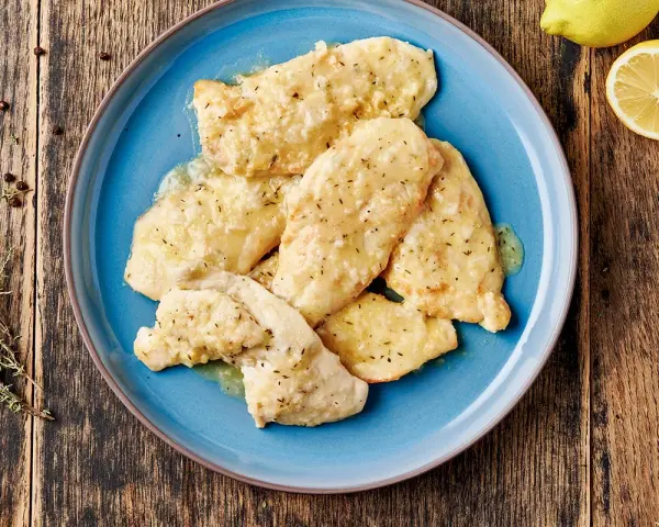 Delizioso petto di pollo in salsa cremosa, guarnito con erbe aromatiche e servito su un piatto blu. Accanto, limone e pepe nero.