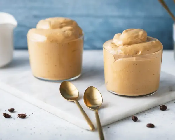 Due bicchieri di cremosa schiuma di caffè, perfetti per una ricetta crema di caffè senza panna. Due cucchiaini dorati e chicchi di caffè completano la scena.