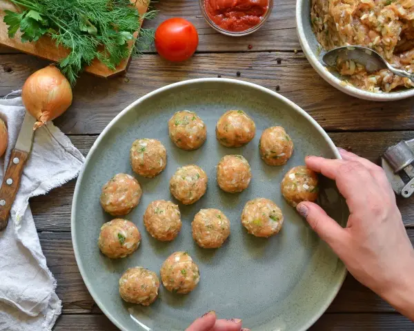 Polpette di pollo pronte per essere cucinate, disposte su un piatto grigio, con ingredienti freschi come cipolle e pomodoro sullo sfondo.