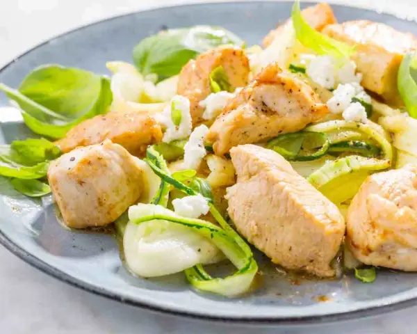 Succulento pollo e zucchine a spirale, guarnito con formaggio sbriciolato e basilico fresco, servito su un piatto blu.