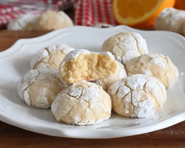 Biscotti alla ricotta soffici e profumati, cosparsi di zucchero a velo, con una fetta d'arancia sullo sfondo.