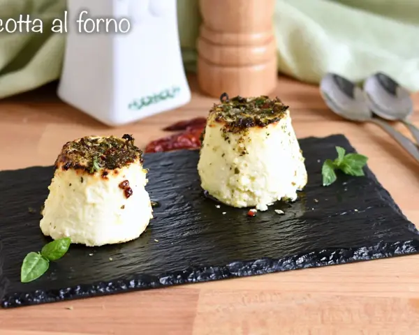 Due tortini di ricotta al forno, dorati e profumati, serviti su ardesia con basilico e peperoncino.