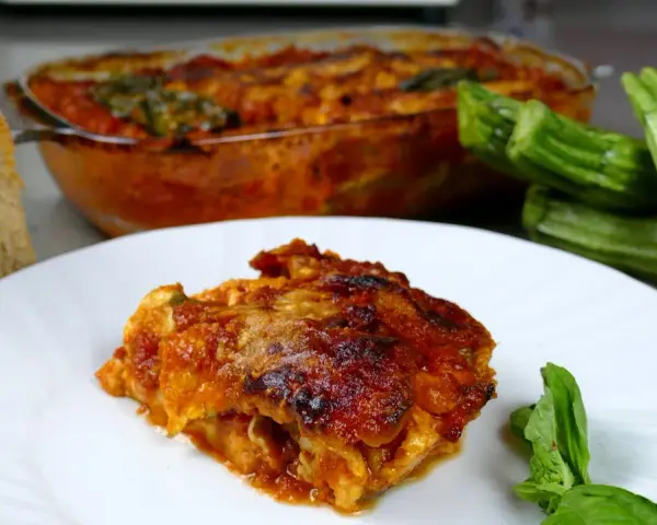 Una fetta di parmigiana di zucchine alla siciliana, dorata e succulenta, accanto a zucchine fresche e pane casereccio.