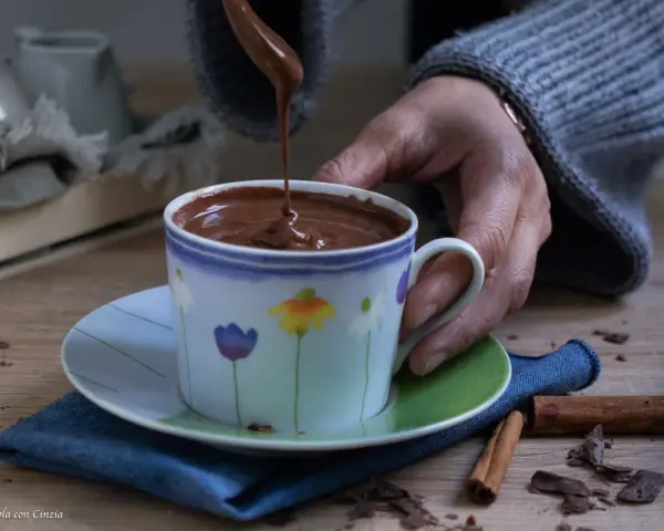 Una mano versa cioccolata fatta in casa in una tazza fiorata. Profumo di cannella e scaglie di cioccolato sul tavolo.