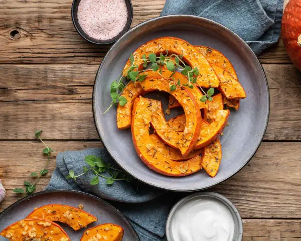 Fette di zucca in agrodolce in padella, condite con erbe aromatiche e servite su un piatto grigio, con contorno di aglio e sale rosa.