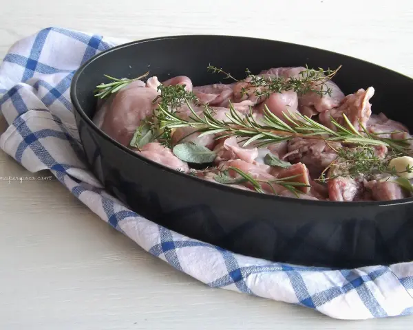 Pezzi di coniglio crudo in una teglia, pronti per essere cucinati al forno con rosmarino, salvia e aglio.