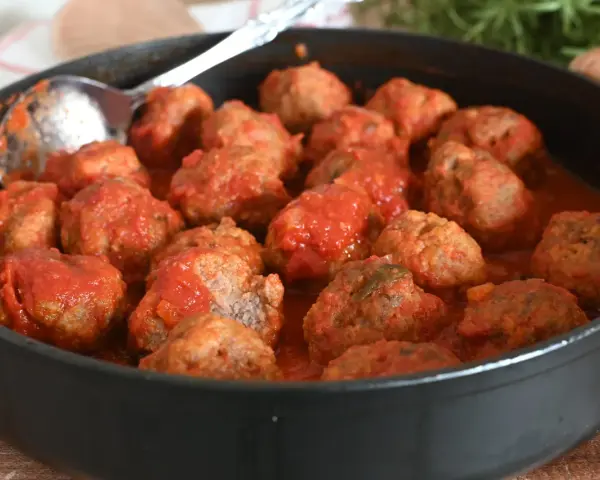 Un tegame pieno di polpette al sugo, pronte per essere gustate. Un cucchiaio d'argento è appoggiato sul bordo, invitando a servirle.