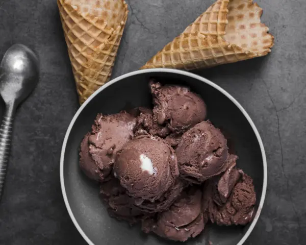 Coppa di **gelato al cioccolato** cremosa, con coni vuoti e un cucchiaio d'argento su sfondo scuro.