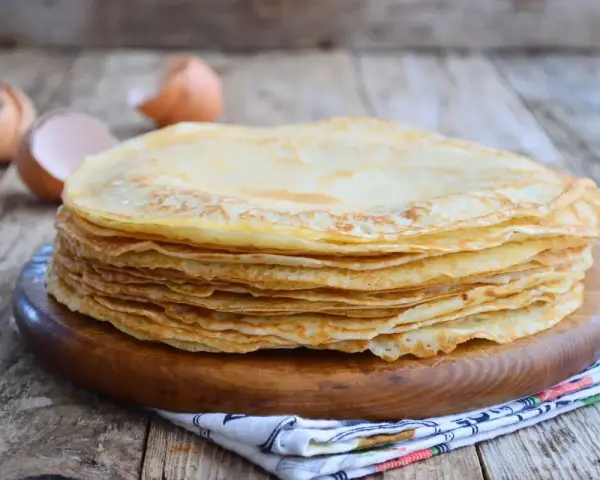 Pila di crepes dorate, perfette per una crepes ricetta senza burro. Guscio d'uovo sullo sfondo.