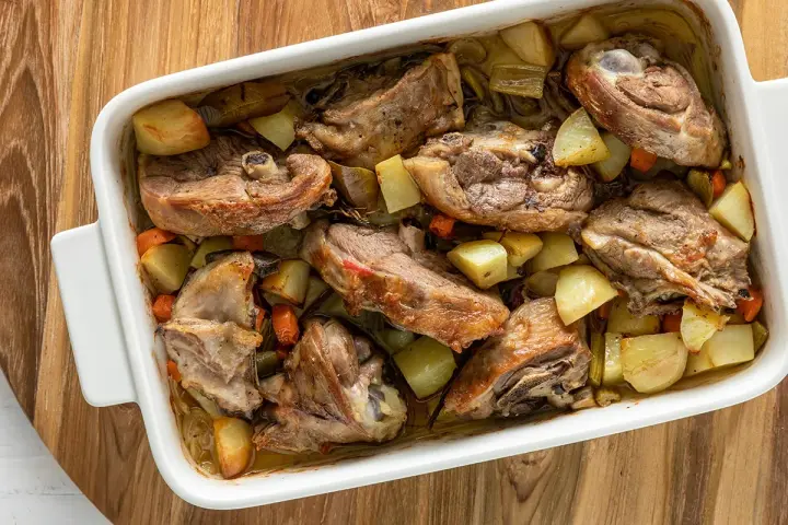 Pezzi succulenti di capretto al forno alla siciliana con patate e carote in una teglia bianca.