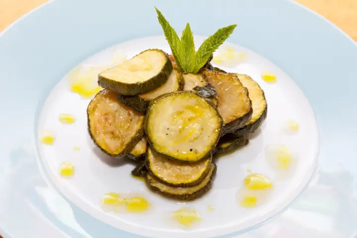 Fette di zucchine alla scapece, condite con olio e guarnite con menta fresca, su un piatto bianco.