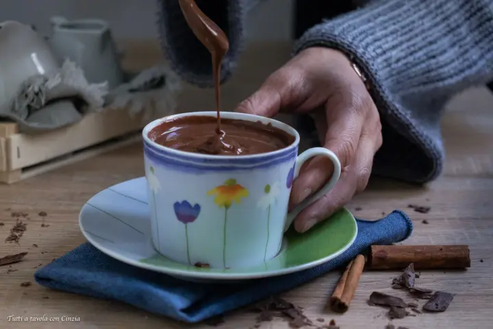 Una mano versa cioccolata fatta in casa in una tazza fiorata. Profumo di cannella e scaglie di cioccolato sul tavolo.