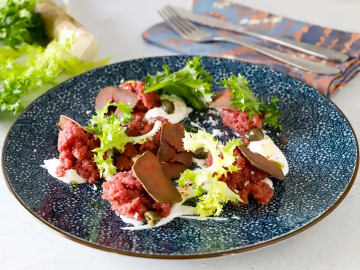 Deliziosa tartare di manzo con lattuga croccante, capperi e una crema bianca su un piatto blu decorato.