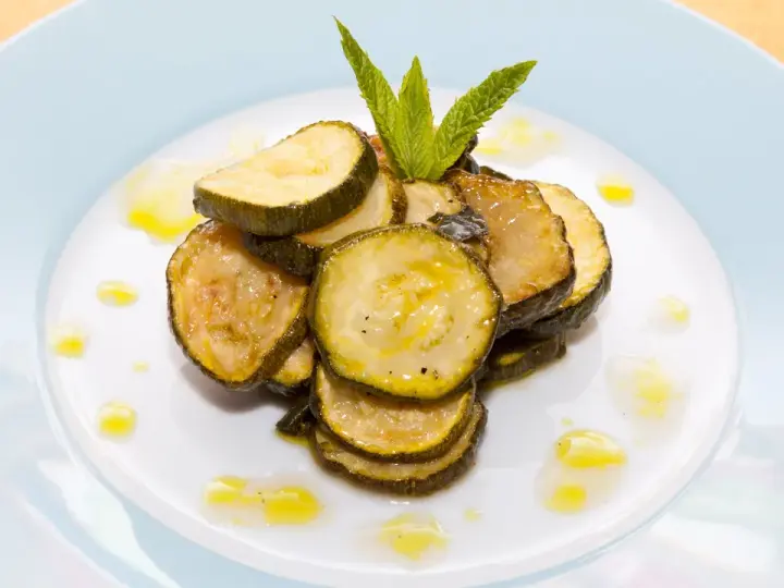 Fette di zucchine alla scapece, condite con olio e guarnite con menta fresca, su un piatto bianco.