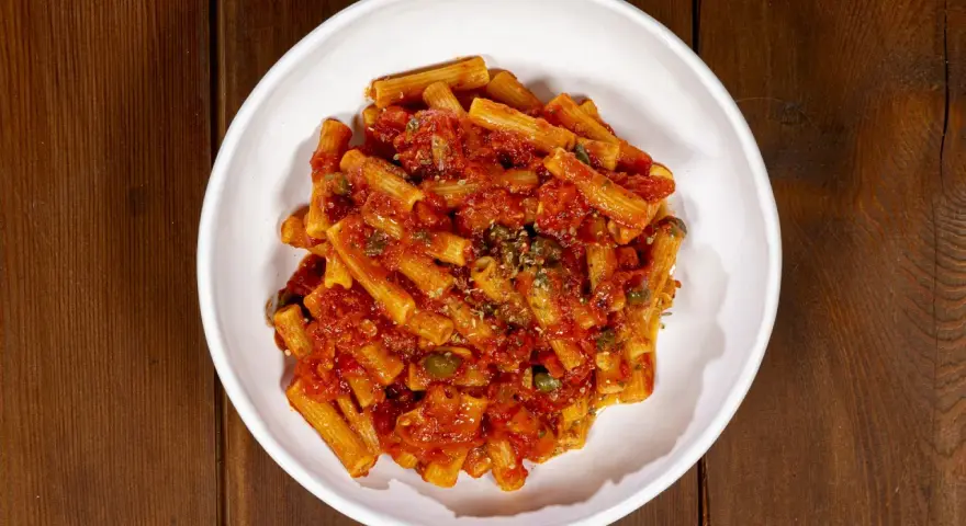 Un piatto di pasta alla pizzaiola, con rigatoni conditi da un sugo rosso ricco di pomodoro, capperi e origano, servito in una ciotola bianca su un tavolo di legno.