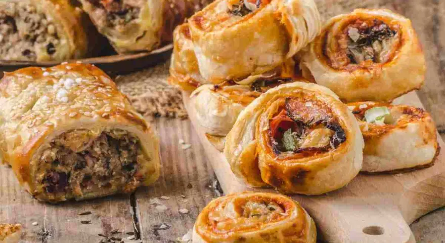 Rustici con pasta sfoglia ripieni di carne e verdure, alcuni cosparsi di semi di sesamo, su un tagliere di legno.