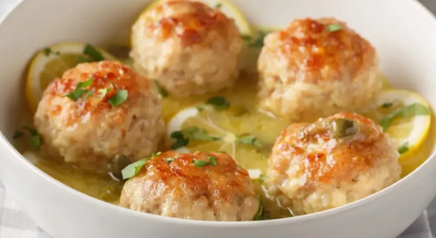 Polpette al limone dorate e succulente, servite in una ciotola bianca con fette di limone e prezzemolo tritato.