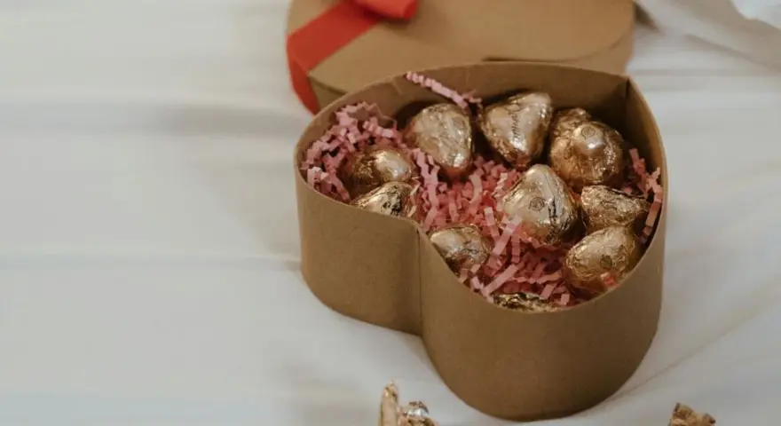 Scatola a forma di cuore con cioccolatini fatti in casa dorati su paglia rosa. Un'altra scatola a forma di cuore con fiocco rosso è sullo sfondo.