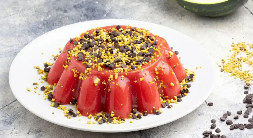Delizioso gelo di melone rosso, guarnito con gocce di cioccolato e pistacchi tritati, su un piatto bianco.