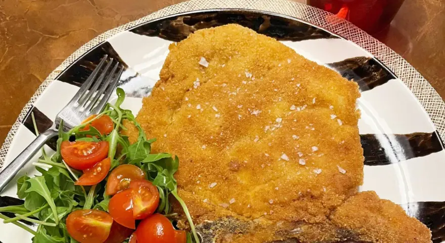 Una dorata cotoletta alla milanese, croccante e cosparsa di sale grosso, servita con un'insalata fresca di rucola e pomodorini.