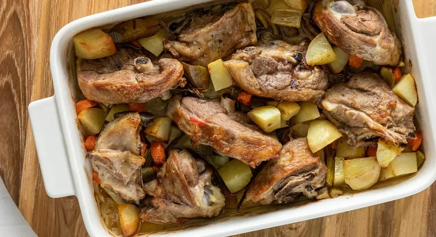 Pezzi succulenti di capretto al forno alla siciliana con patate e carote in una teglia bianca.