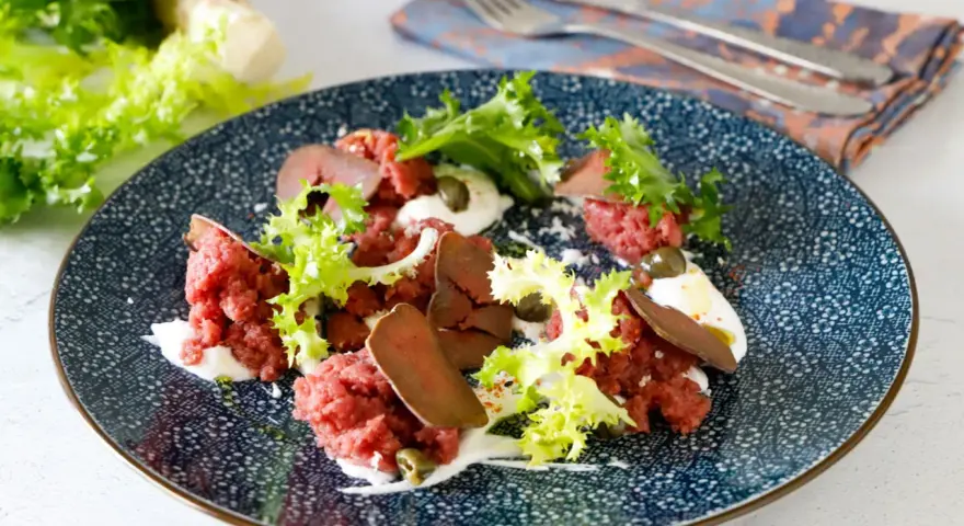 Deliziosa tartare di manzo con lattuga croccante, capperi e una crema bianca su un piatto blu decorato.