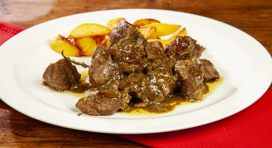 Un saporito abbacchio alla cacciatora, tenero e succulento, servito con patate arrosto su un piatto bianco.