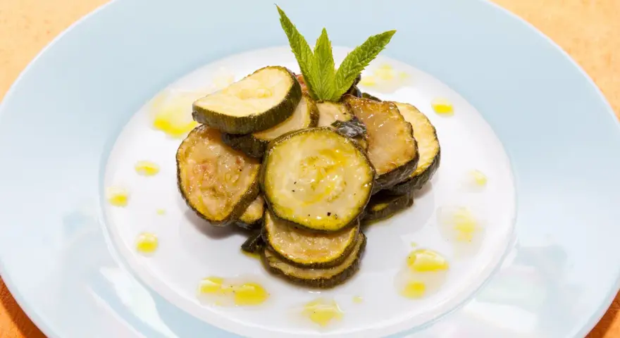 Fette di zucchine alla scapece, condite con olio e guarnite con menta fresca, su un piatto bianco.