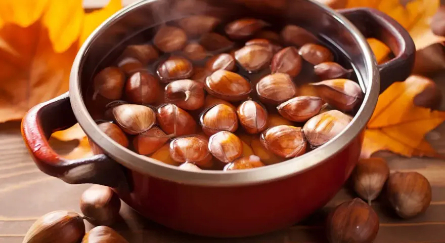 Castagne bollite con buccia in una pentola rossa, circondate da foglie autunnali.